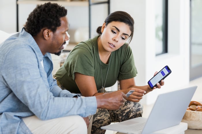 Two people are in a living room while one holds a phone and the other holds up a credit card. 