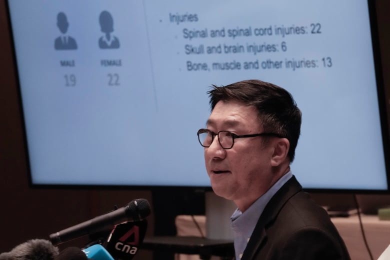 A man with black hair, black glasses and a black suit stands at a podium during a presentation for reporters.