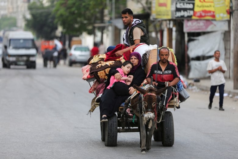 Three adults and a child are shown riding on a cart that is being led by an unspecified animal, on a paved road with buildings and onlookers shown in the background.