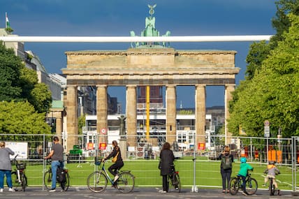 Berlin, Mai 2024, UEFA EURO 2024, hier entsteht die größte Fan Zone Deutschlands für gemeinsames Public Viewing und Feiern, auf der Straße des 17. Juni wurde spezieller Kunstrasen ausgelegt, Berliner und Touristen bleiben neugierig am Bauzaun stehen, im Hintergrund das Brandenburger Tor, direkt davor das größte Fußballtor der Welt, hier zu sehen ein weißer Balken, die Seitenpfosten sind durch Bäume verdeckt.