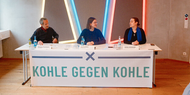 Drei Personen sitzen bei einer Pressekonferenz an einem Tisch. Davor ist ein Plakat gespannt mit dem Spruch: Kohle gegen Kohle