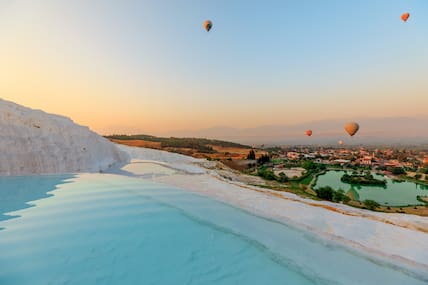 Kalksteinfelsen mit Wasserbecken in Pamukkale, im Hintergrund eine wasserreiche Landschaft, über der Heißtluftballons schweben