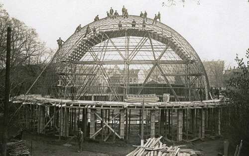 Black and white photo of a domed structure under construction with workers standing on the top.