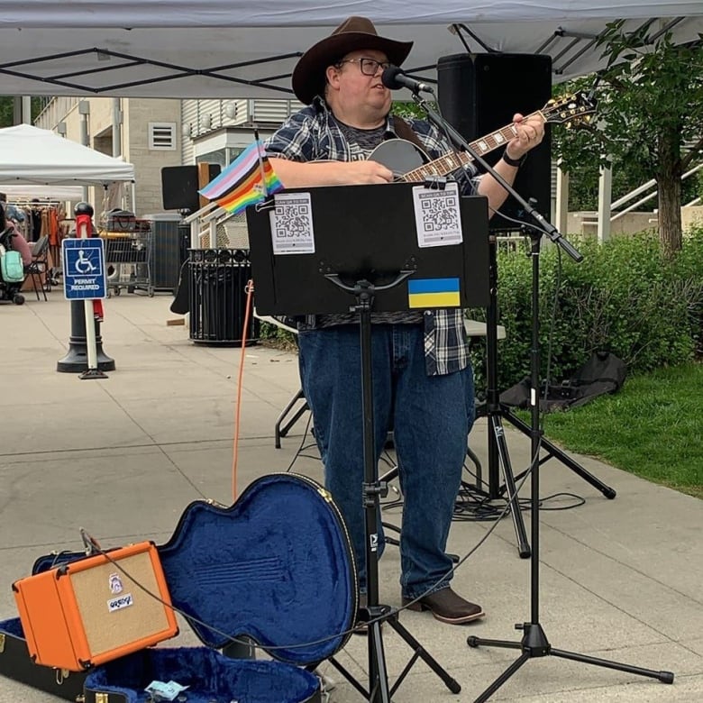 A man plays guitar in an outdoor setting. He is wearing a cowboy hat.