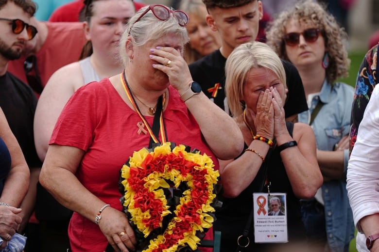 Two women stand outside in a crowd, crying and holding their hands over their faces. 