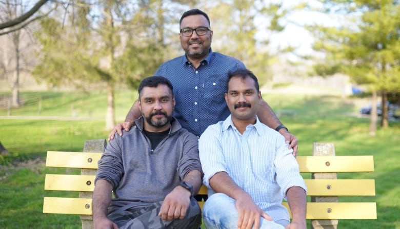 Three South Asian men post on a park bench outdoors.