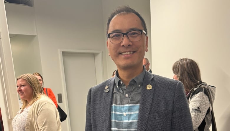 Vertical photo of a middle-aged Asian-Canadian man, wearing a blazer and jeans. Indoor photo. Several people in the background. 