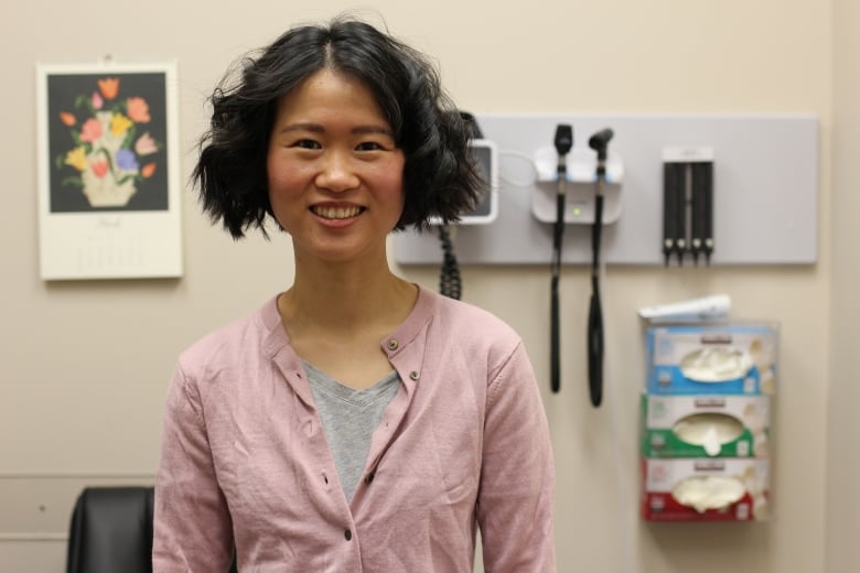 A woman with a black bob, a grey t-shirt and pink cardigan is pictured standing inside a patient exam room at a family medicine clinic.