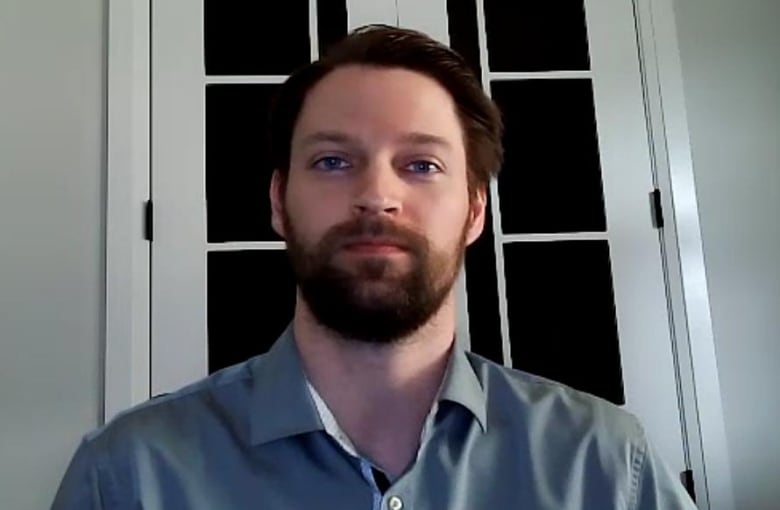 Redel has a beard and is wearing a grey dress shirt as he sits in front of a white door.