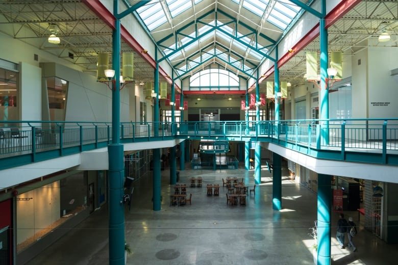 An empty large building with tables and chairs in the middle of it. there are skylights and two people walking through the large open space.