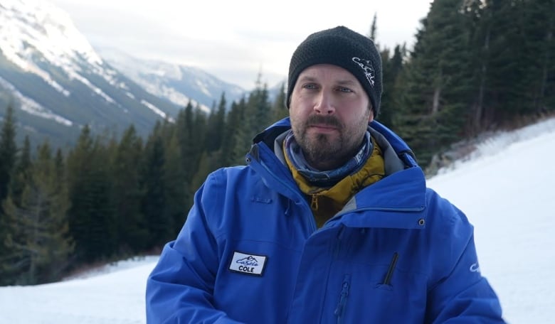A man in a black toque and blue jacket stands in front of mountains. 