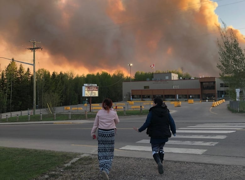 Two people walk toward a school as smoke rises in the background.