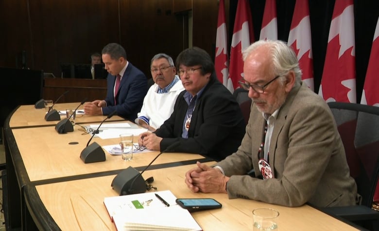 A group of men sitting behind a long desk with Canadian flags in the background.