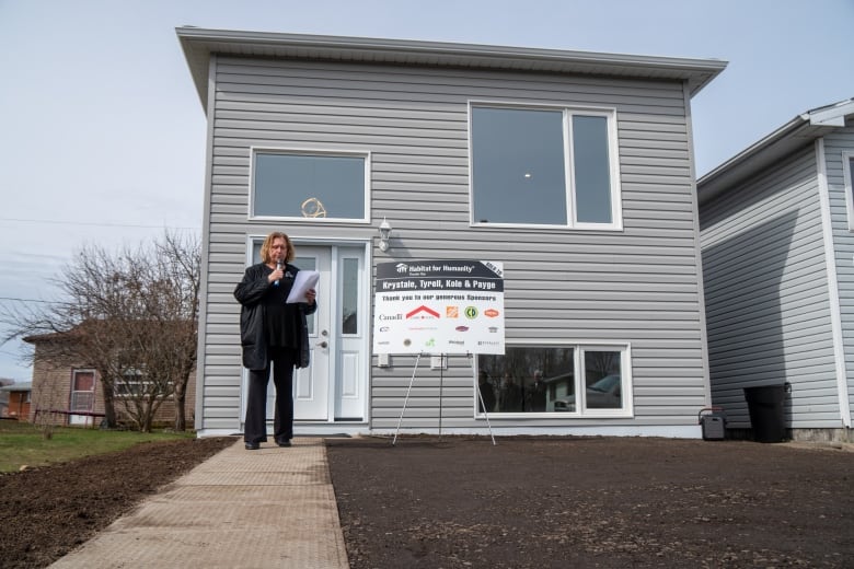 A wide shot of a small, light grey home. A person stands on the walkway and speaks into a microphone.