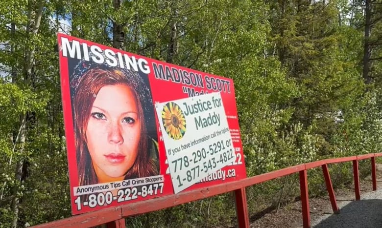 Surrounded by trees, a red missing persons billboard shows the face of a young woman, her name, and the number to call to provide anonymous tips to Crime Stoppers.  