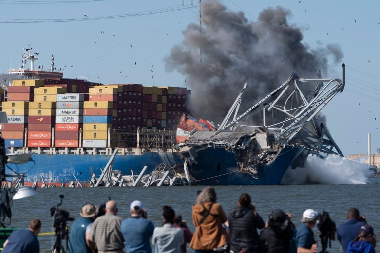 Several people with their backs to the camera are shown on land in the foreground as they watch smoke rise from a large ship that contains containers and has bridge wreckage draped atop it.