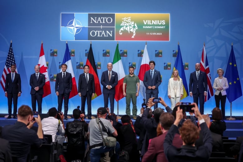 Ten G7 leaders stand on stage with European leaders and Ukrainian President Volodomyr Zelenskyy in front of flags of various countries. 