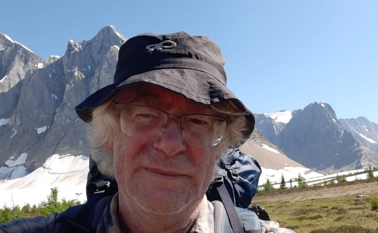 A man wearing a hat looks at the camera with a backdrop of mountains pictured behind him.