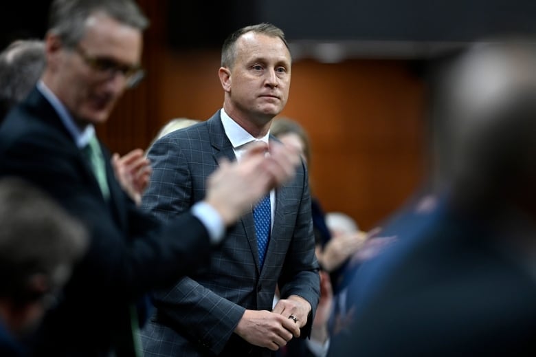 A man in a suit and tie stands in the House of Commons. Others around him applaud.