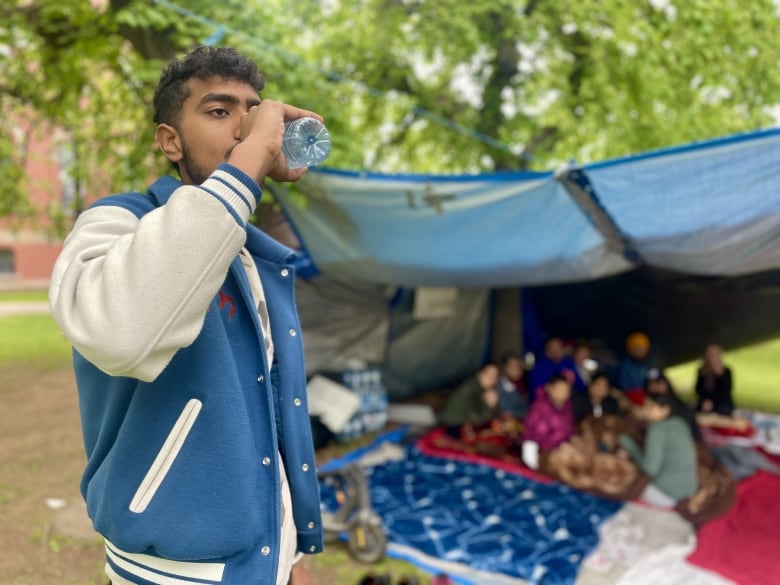 A man in a blue and white jacket with short drak hair sips from a plastic water bottle in front of an encampment.