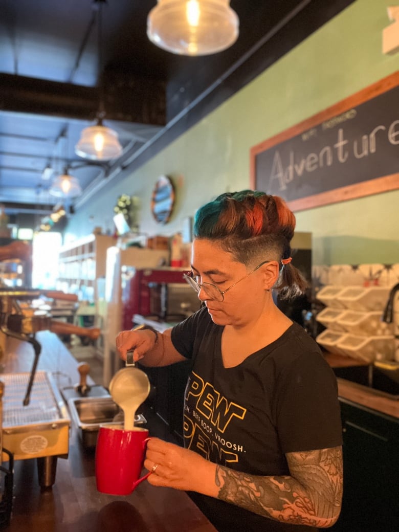 A Caucasian woman wearing a black Star Wars t-shirt pours a latte while working at a cafe.