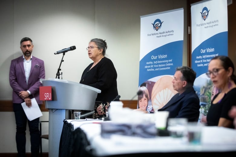 An Indigenous woman speaks in front of two banners reading 'First Nations Health Authority'. She is flanked by other people.