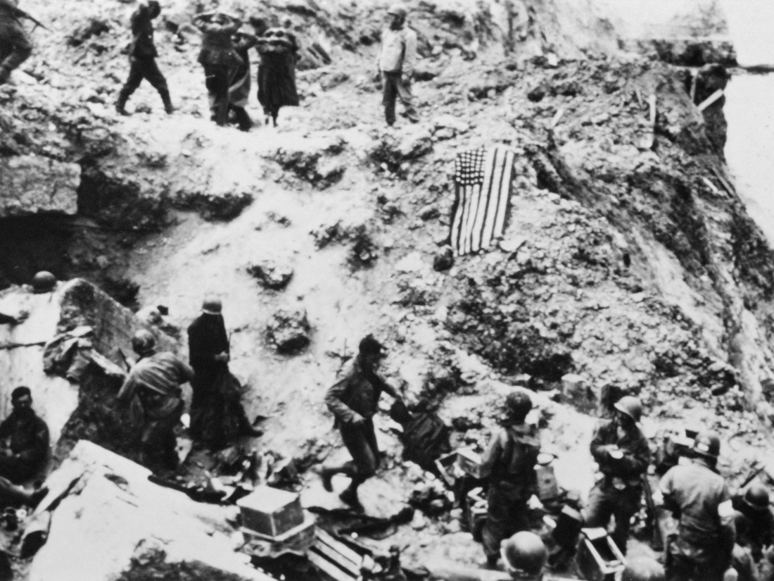 Soldiers and an American flag stand on a rocky cliffside.