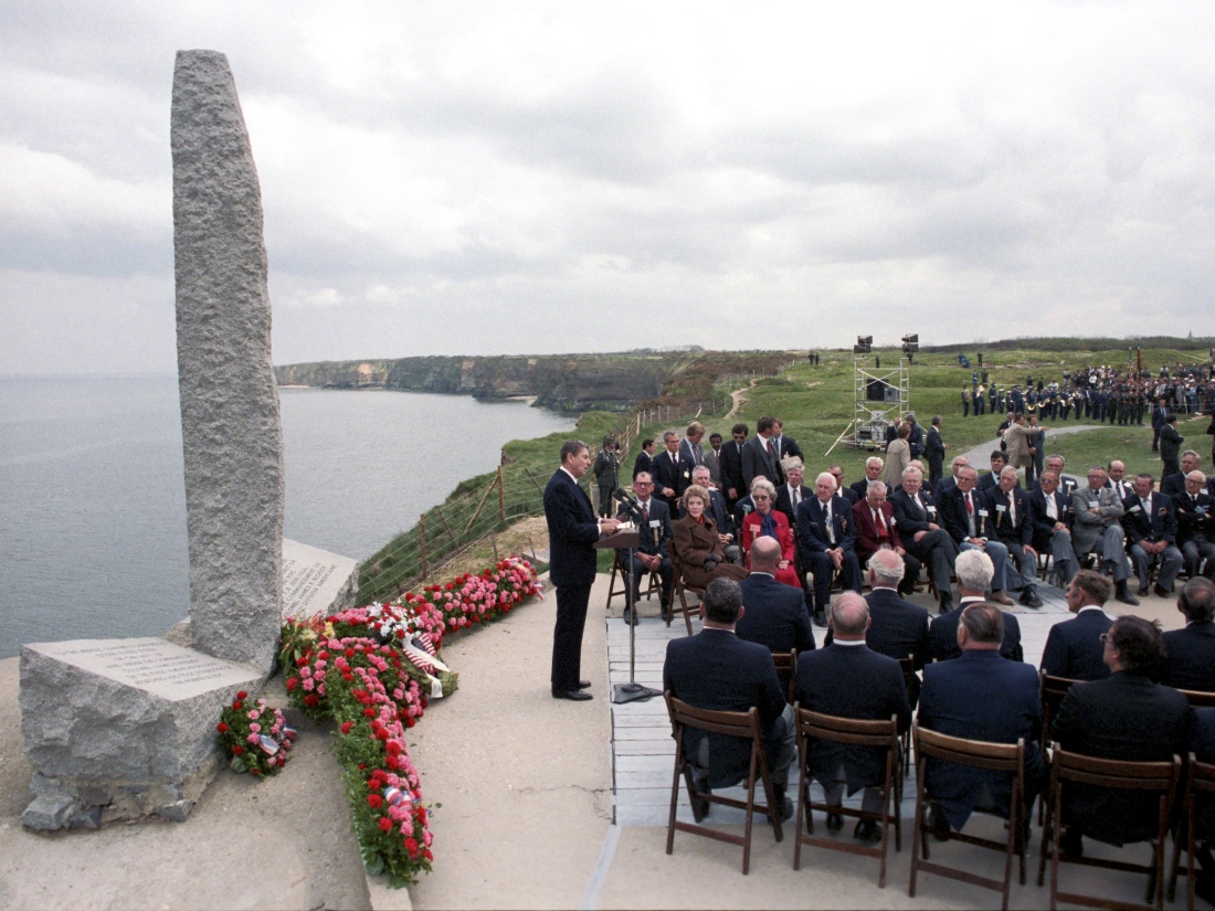 A crowd of mostly men listens as Ronald Reagan speaks at a podium in front of a granite monument on a clifftop overlooking the water.