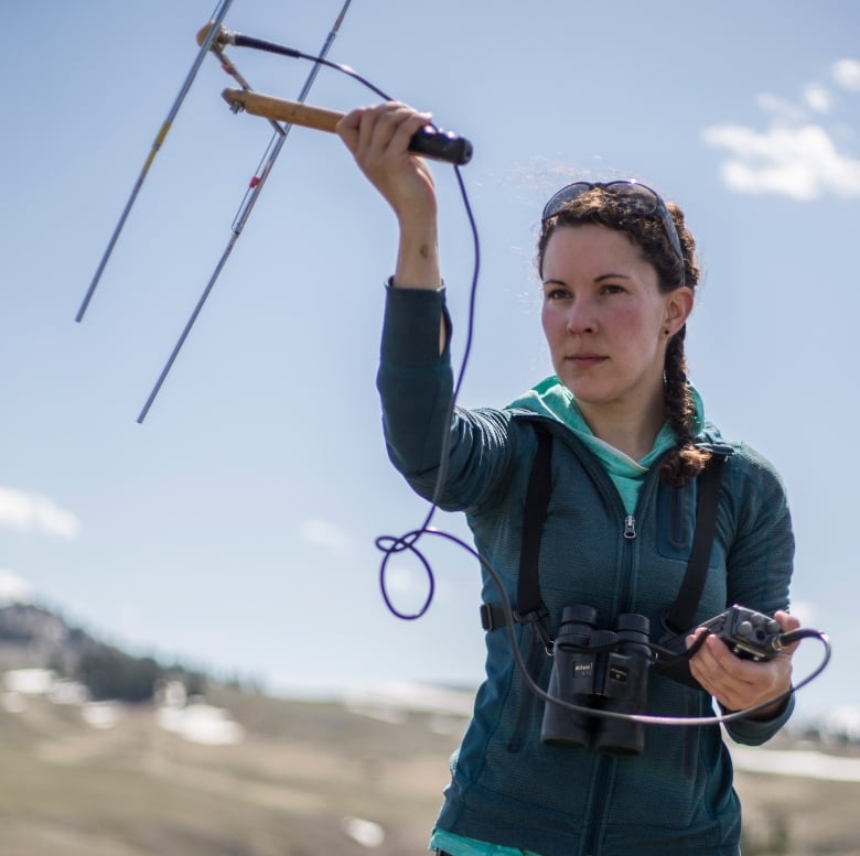 A woman, her hair braided over one shoulder, stands outside holding an antenna in the air attached by a cord to a hand-held device. She's wearing sporty attire and has a pair of binoculars around her neck. 