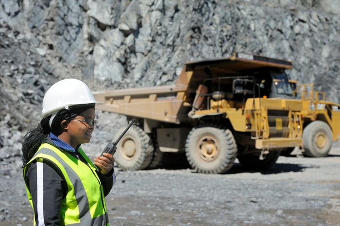 A person in protective gear talking on a communications device with a dump truck in the background.