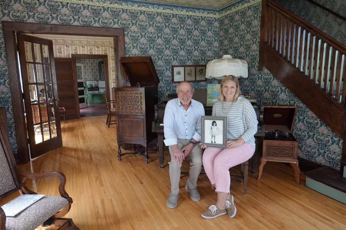 Two people sitting with photo: John Kelsch, left, and Janie Heitz, right, holding a framed photo of a young Judy Garland. They are the former and current directors of the Judy Garland Museum in Grand Rapids, Minnesota. They posed for a photo in the actress’ childhood home on May 30.