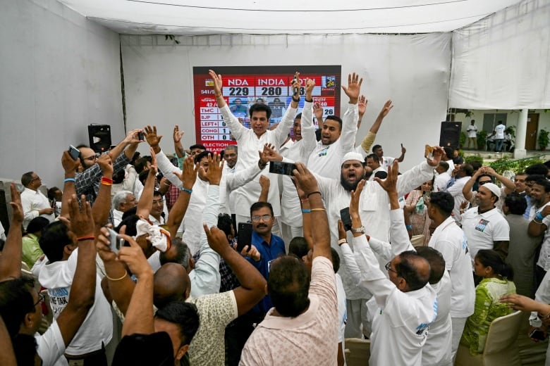 Several people wearing white raise their arms inside a room where a television is being played in the background.