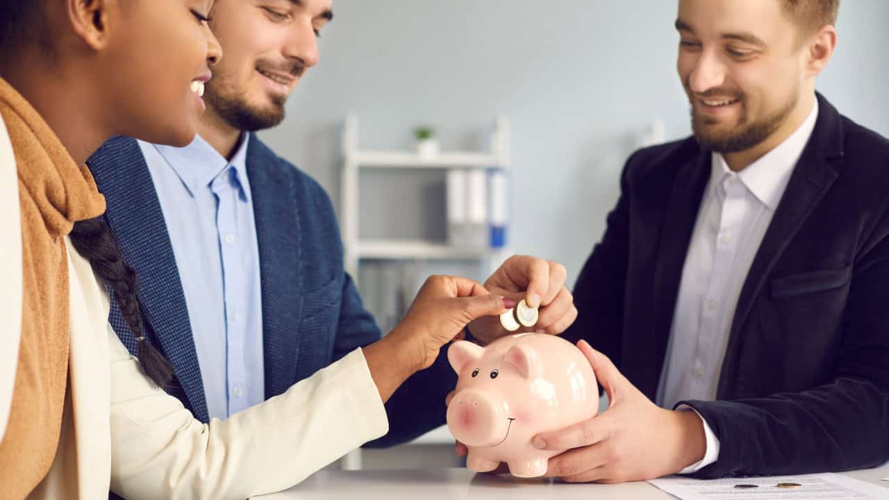 Happy interracial couple putting coin into piggy bank on meeting with advisor