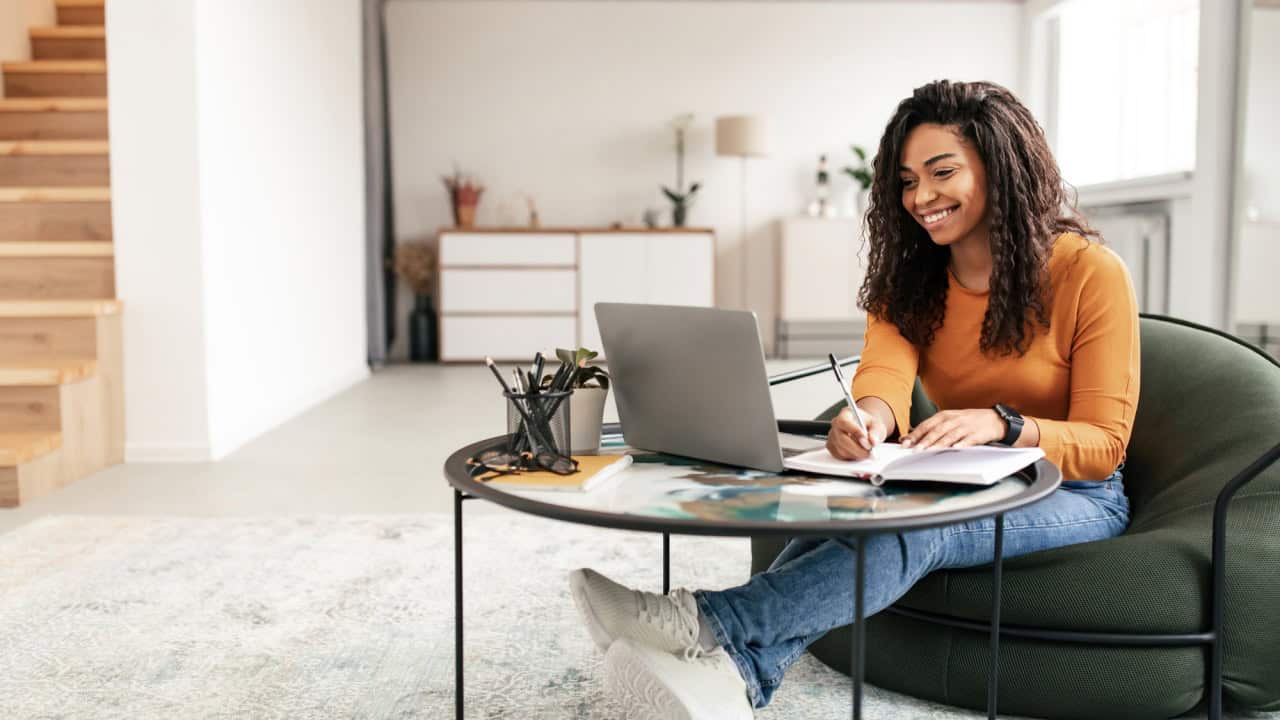 Smiling woman sitting at table, using computer writing in notebook