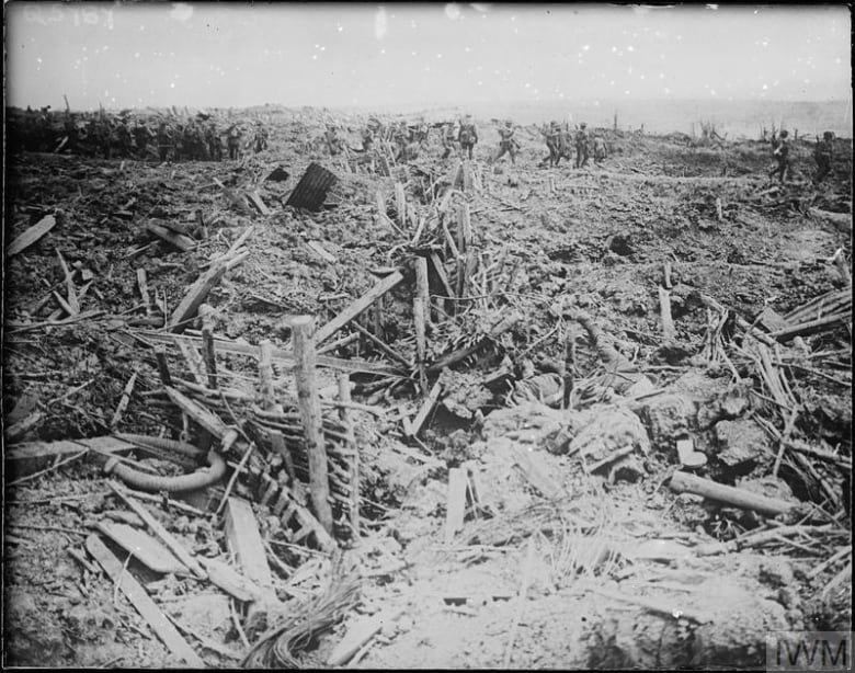 While troops walk in the background in the foreground there is a tangle of splintered wood and cratered ground.