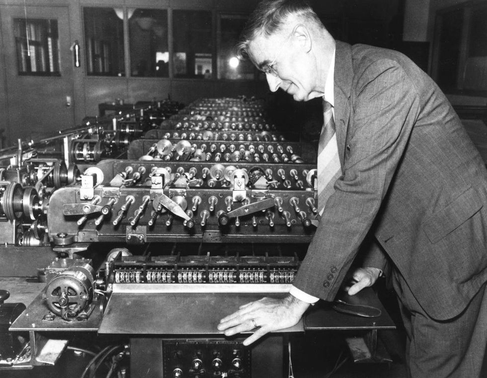 Black and white photo of a man in a suit leaning over a table-length machine with many rows of metal gears, shafts, and cranks.