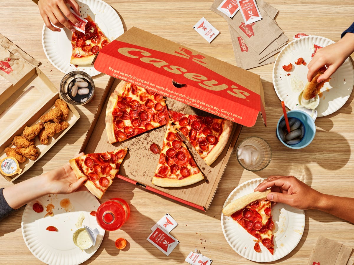 Casey's pepperoni pizza with a box, beverages, various condiments and the hands of people eating.