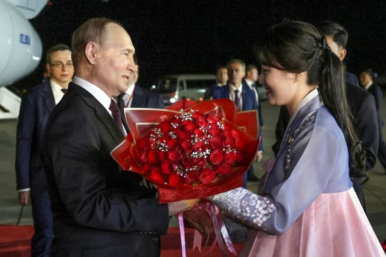 An older balding Caucasian man in a suit and tie is shown in an nighttime photo on an airport tarmac receiving a bouquet of flowers from an Asian woman in a traditional costume.