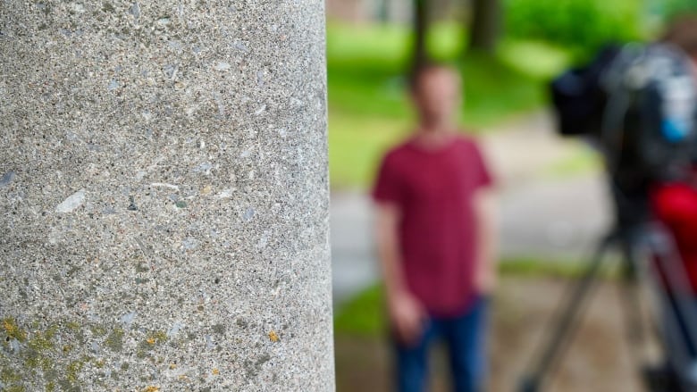 A man wearing a striped shirt and jeans gives an interview. He's out of focus for privacy reasons.