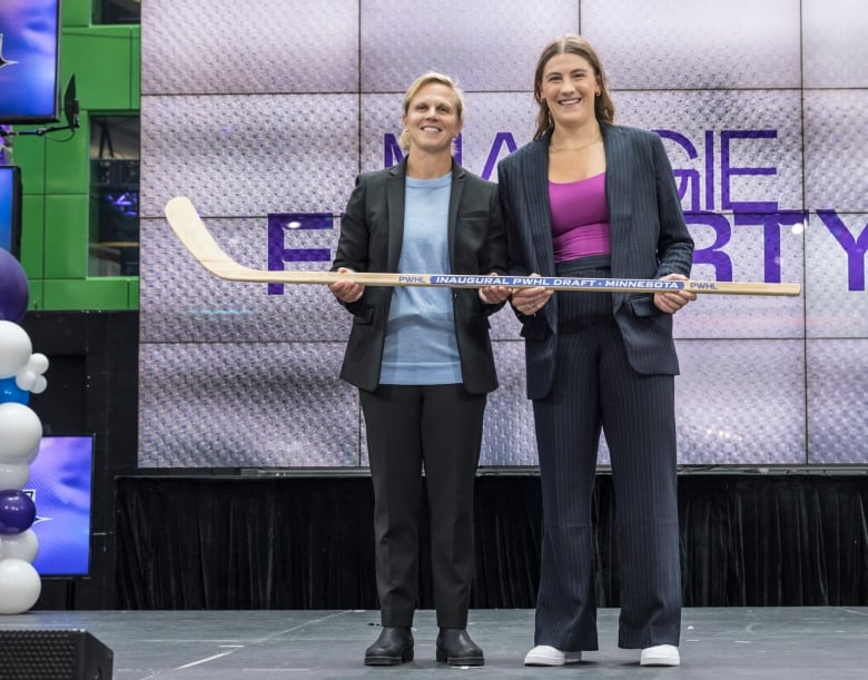 Two women stand on a stage smiling and holding a hockey stick.