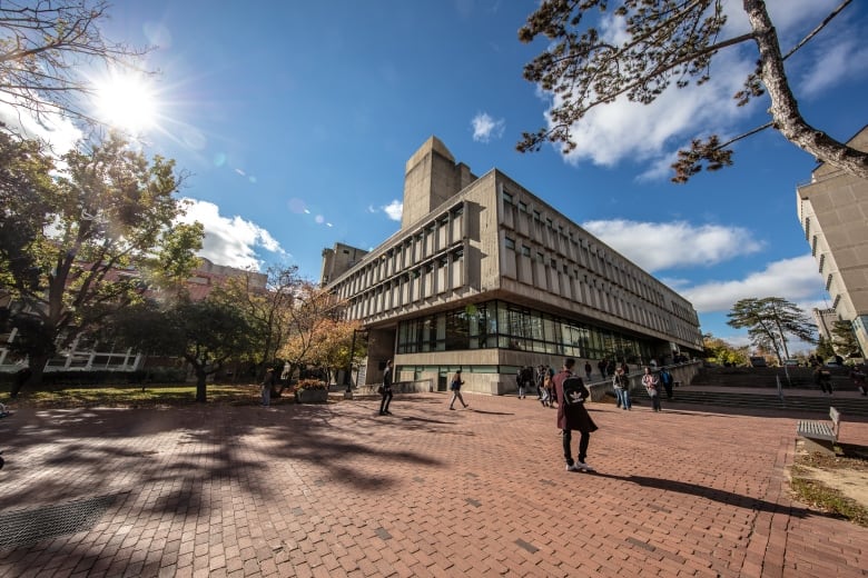 A student carrying a backpack walks in front of a brutalist concrete building under blue sky. 