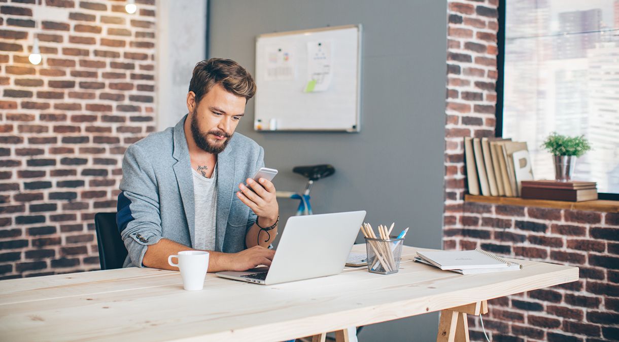 Casual professional man using his computer while swiping through his phone