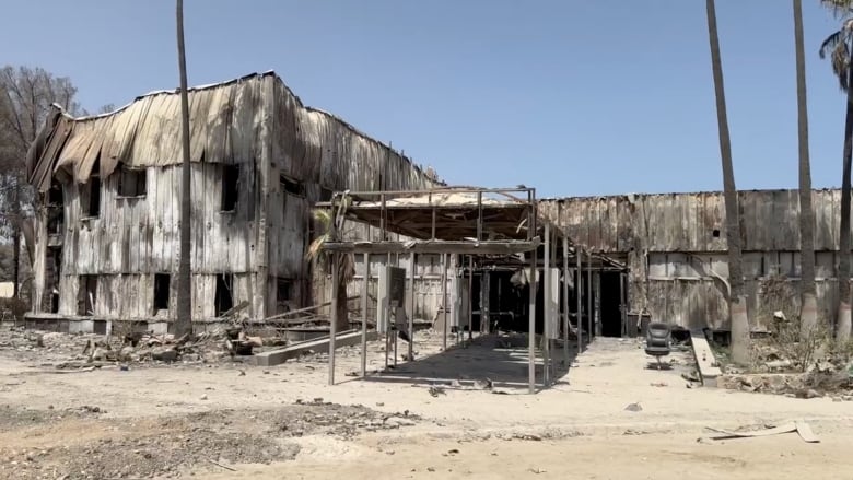 Burned-out buildings on a dirt ground are shown in a daytime photo.