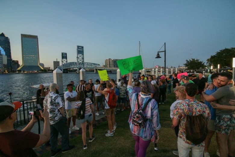 A crowd of people organize on the grass along a riverbank at dusk, with a tall bridge and skyscrapers in the background. 