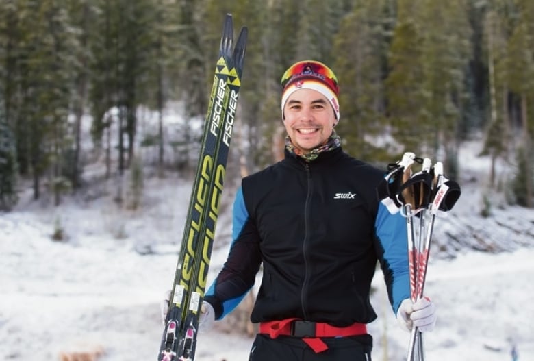 A man smiles at the camera on a snowy cross country ski trail holing his poles and skis. 