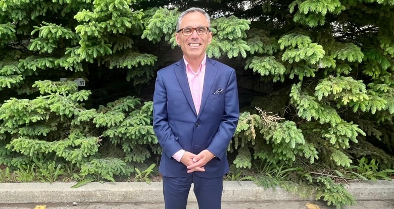 A man in a lue suit stands in front of a row of fir trees.