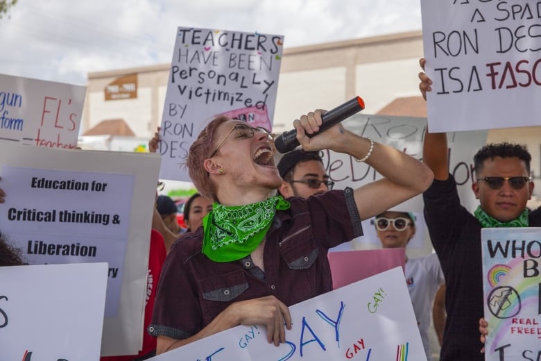 A person with pink-dyed hair, wearing glasses and a green handkerchief around his neck, shouts into a microphone while standing in front of a crowd of protesters. 