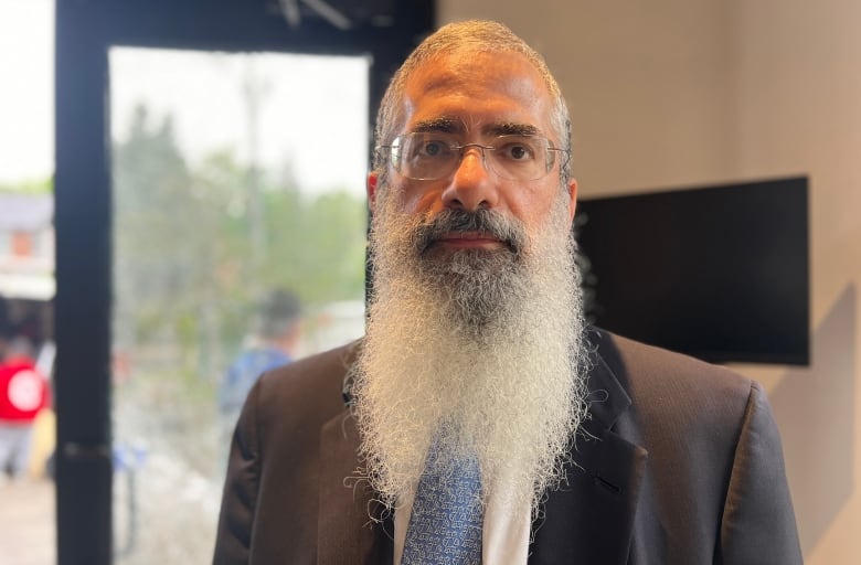 A bearded man in glasses and a suit stands indoors, looking off camera.