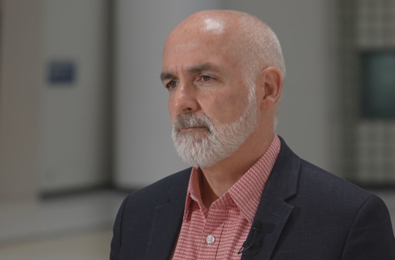 A balding man with a grey beard wearing a suit jacket and red shirt looks to the left.